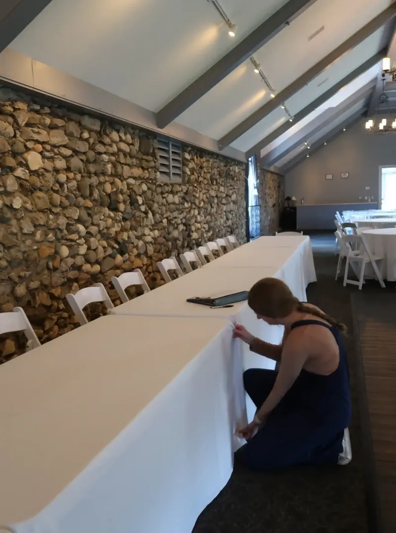 A woman straightens a white tablecloth on a long table in an event space at Castle Farms. 