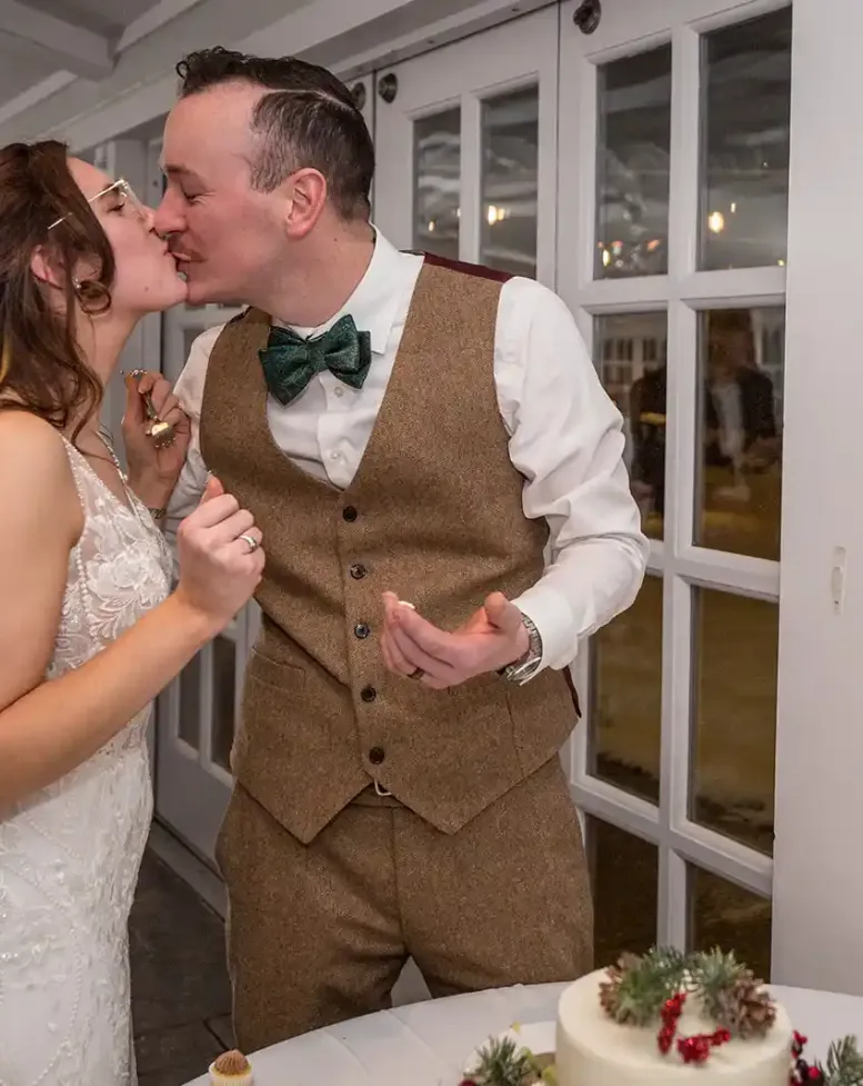 A bride and groom kissing beside their wedding cake next to windows of a reception hall at Castle Farms. 