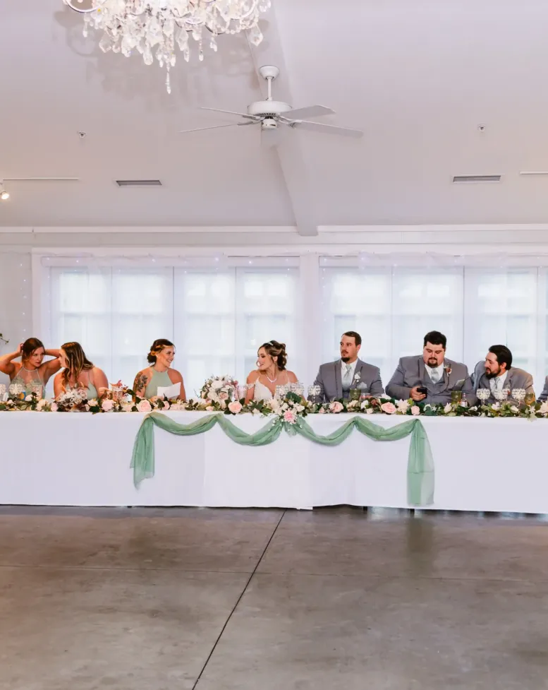 A wedding party sitting at a long table decorated with flowers in a bright reception hall at Castle Farms.