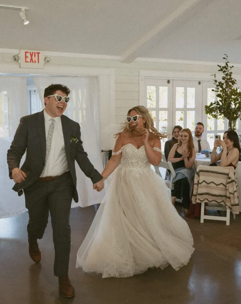 A bride and groom wearing heart-shaped  sunglasses enter a white-walled event space at Castle Farms. 
