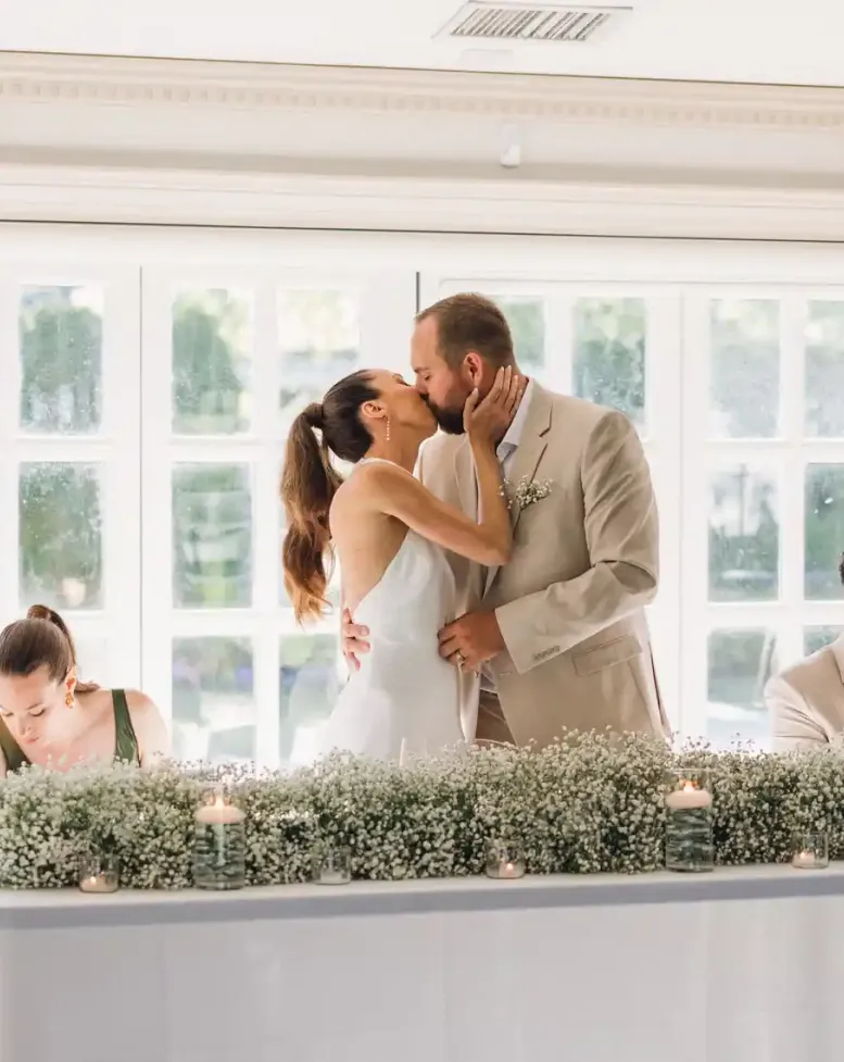 A bride and groom standing at the wedding party table kissing in front of windows in an event space at Castle Farm.