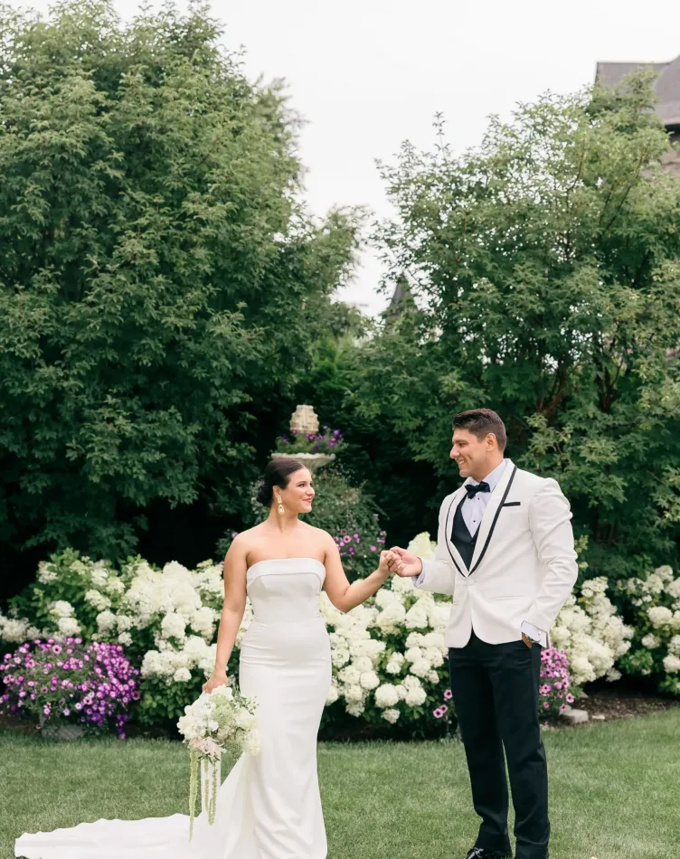 A bride and groom holding hands in a green garden with white flowers at Castle Farms.