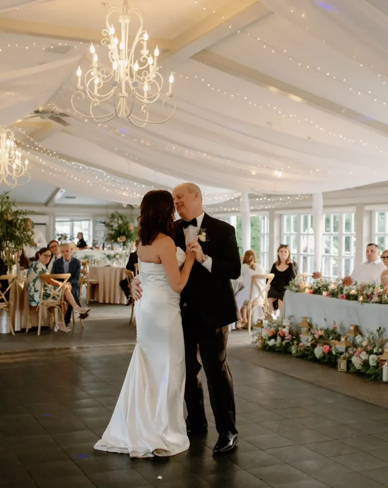 A slow dance with the bride on the dance floor under a golden chandelier with wedding guests watching in an event space at Castle Farms.