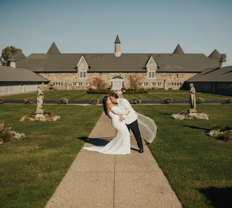 Wedding couple sharing a dip kiss in the Castle Farms formal garden with the stone venue behind them. Photo credit: Zoe Fortuna.