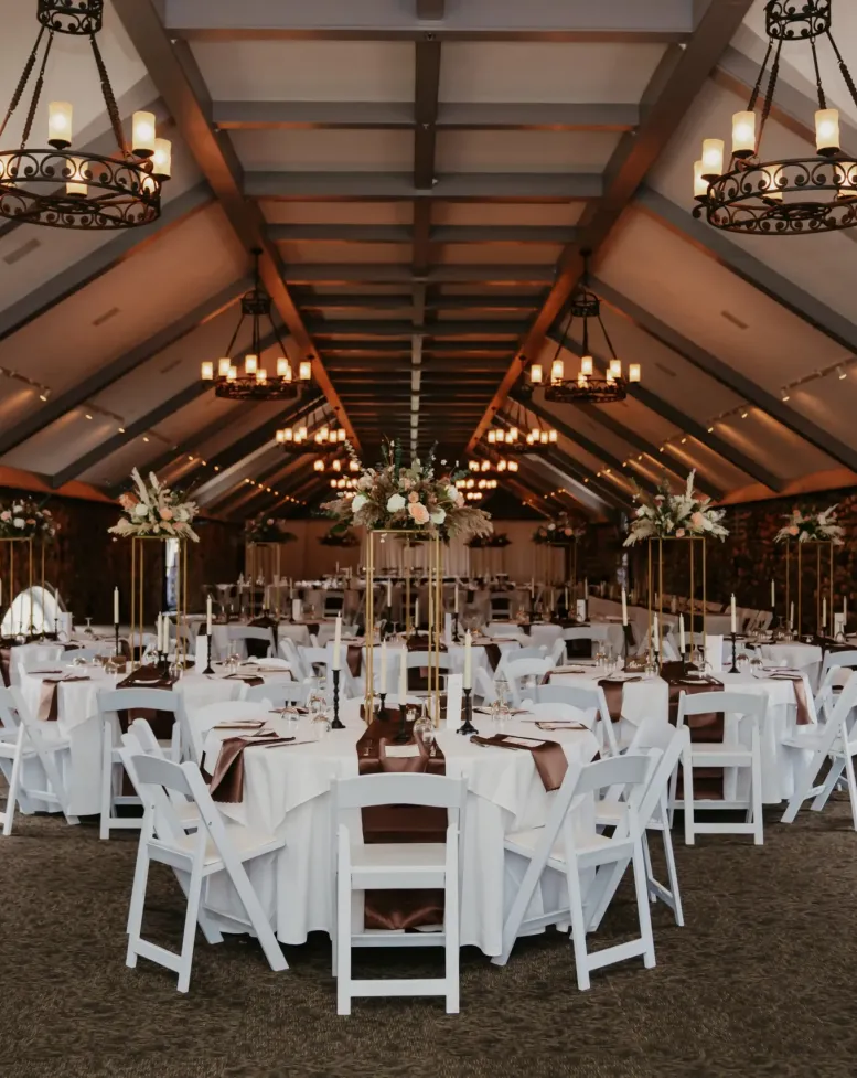 A long stone-walled event space with metal chandeliers and white chairs and round tables at Castle Farms.