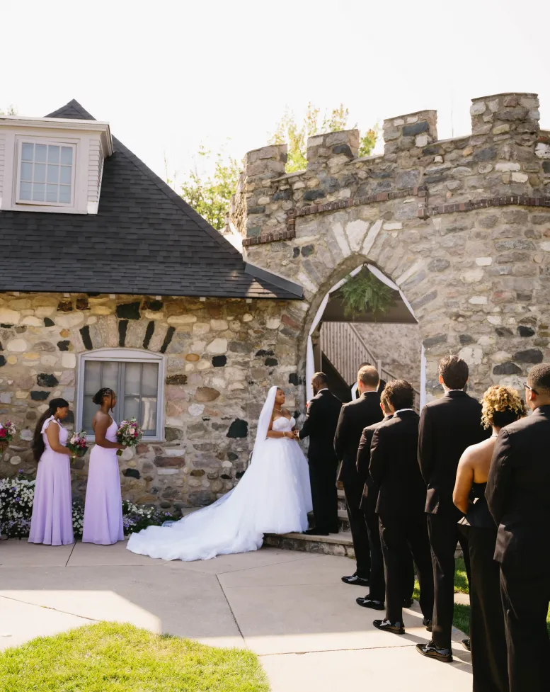 Groomsmen and bridesmaids in a line facing a bride and groom getting married in front of a stone tower at Castle Farms.  