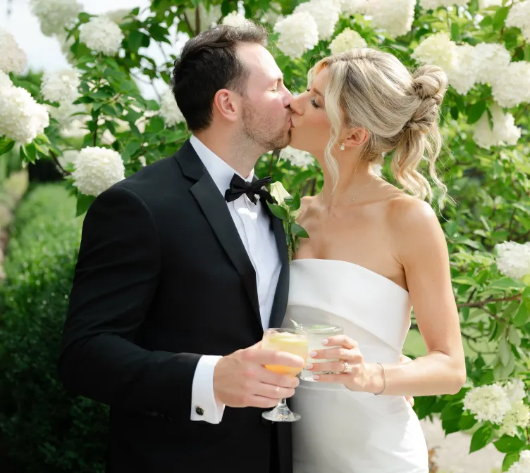 A bride and groom kiss while holding drink glasses in front of flowers at Castle Farms.