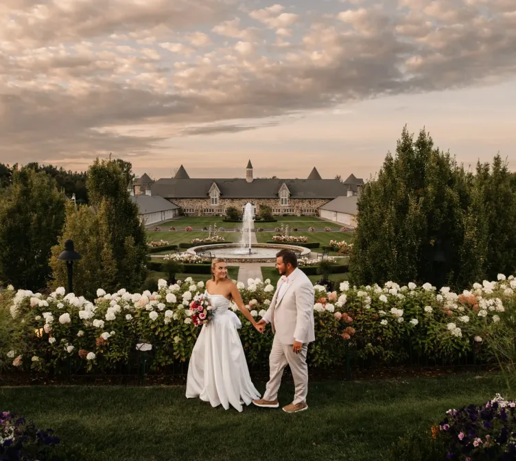 A bride and groom walking by flowers on a golden evening in front of the landscape of Castle Farms.