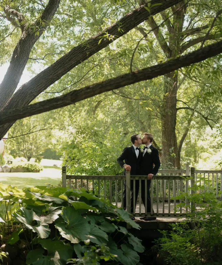 Grooms kiss on a wooden bridge in the shade of green trees at Castle Farms.