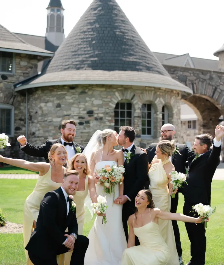 Bride and groom kissing with their wedding party celebrating in front of a Castle Farms stone turret. Photo credit: Sidney Bischer Photography.