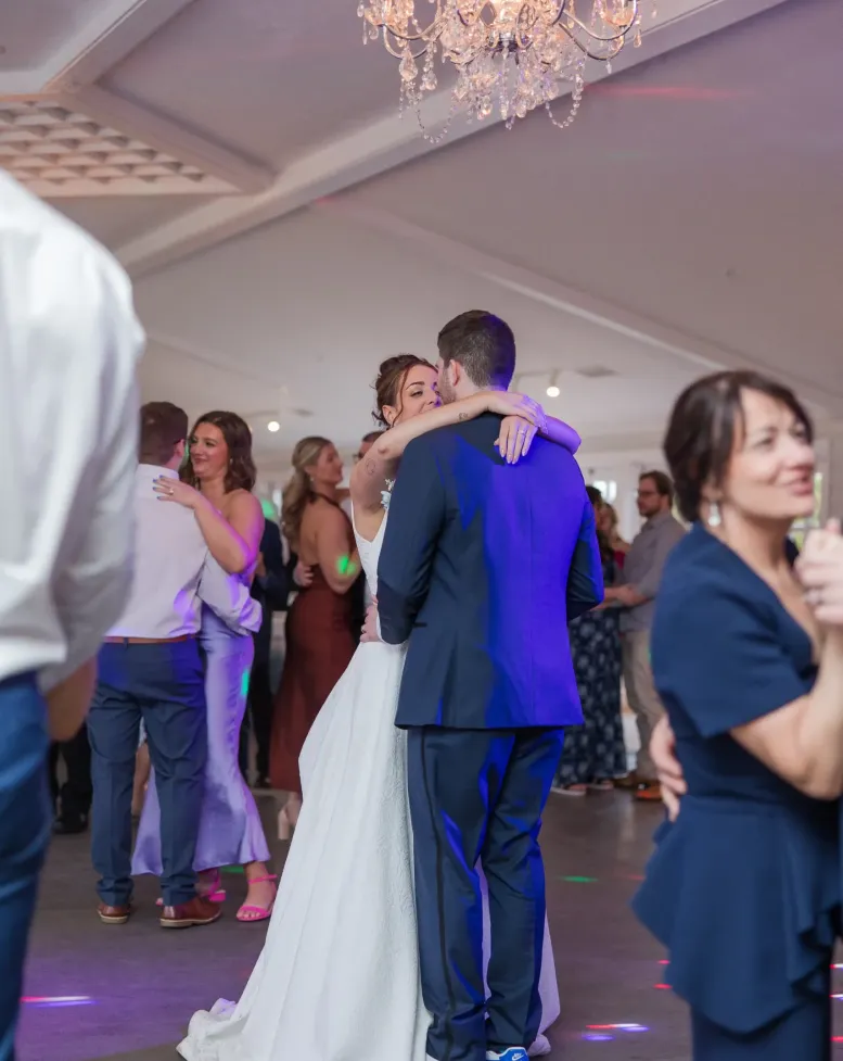 A bride and groom slow dancing surrounded by guests on the dance floor  under a chandelier in an event space at Castle Farms.