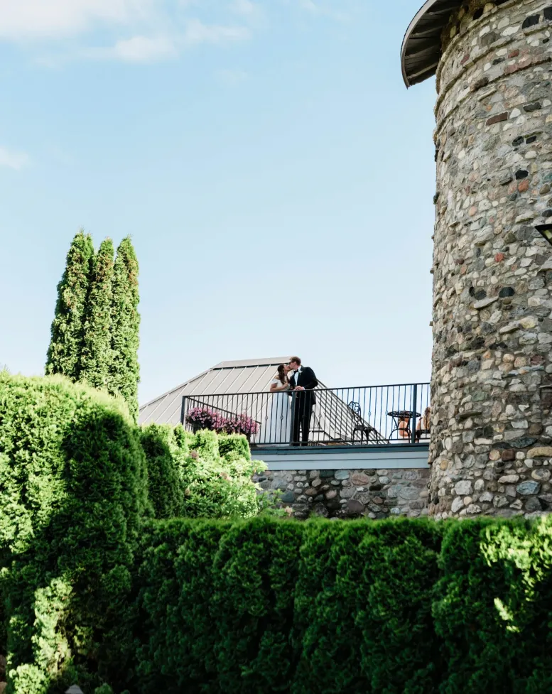 A bride and groom embrace on a balcony beside a tower and hedges under a blue sky at Castle Farms.