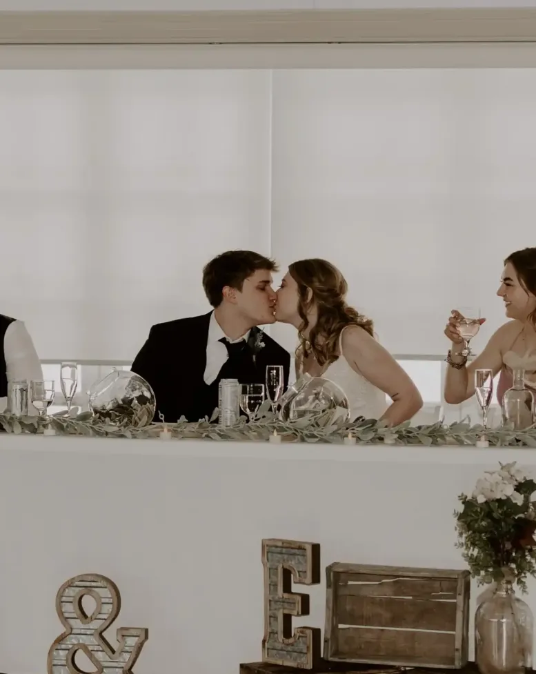 A bride and groom kissing at the wedding party table in front of windows in a reception hall at Castle Farms.