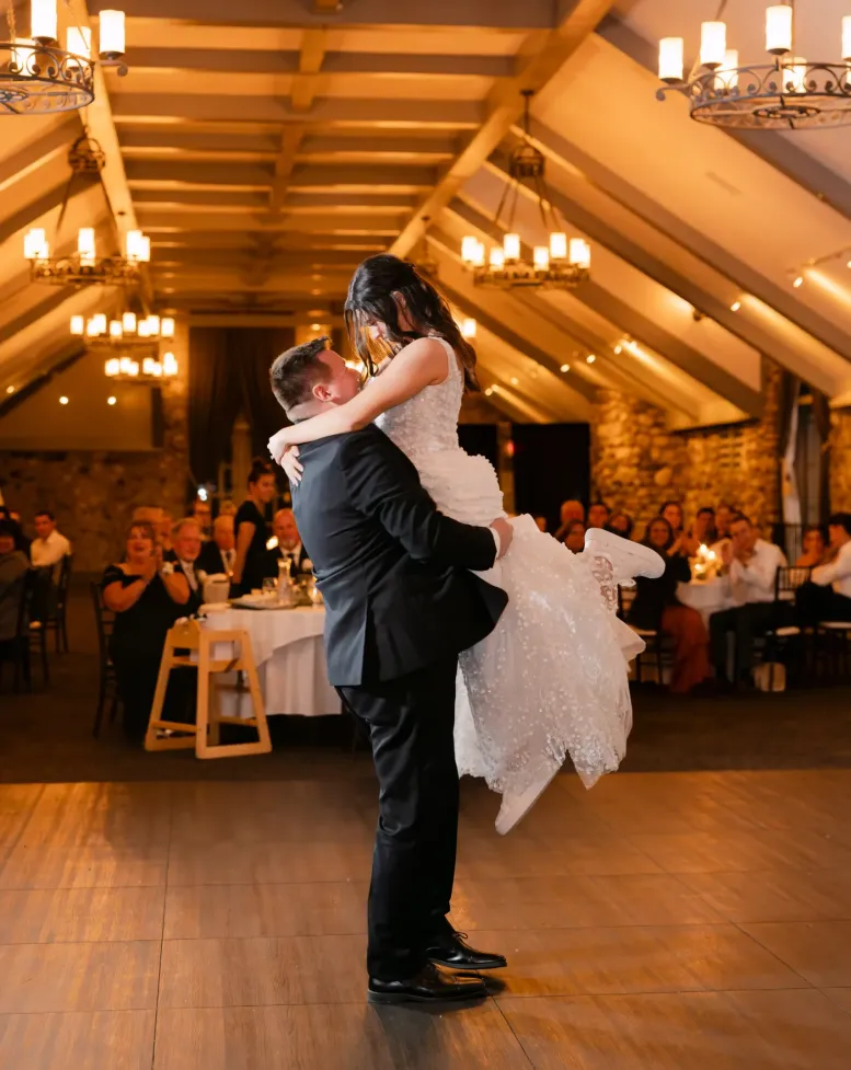 A groom picking up his bride on the dance floor of a golden-lit event space at Castle Farms.