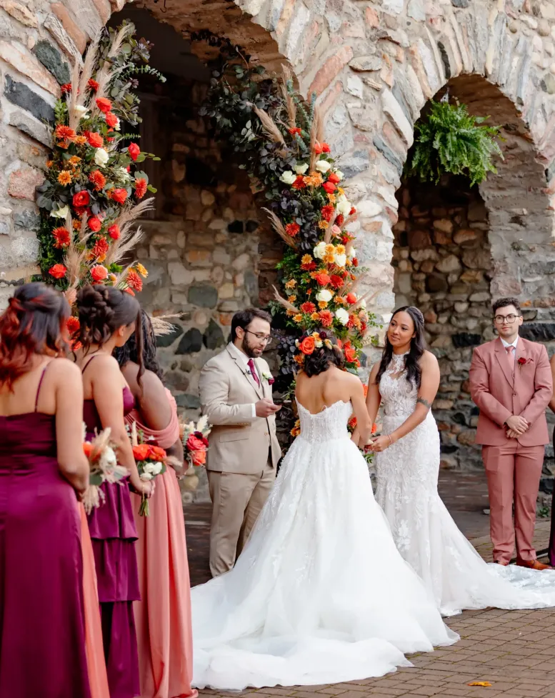Brides at a wedding joined by tropical flowers and bridesmaids in a courtyard at Castle Farms.
