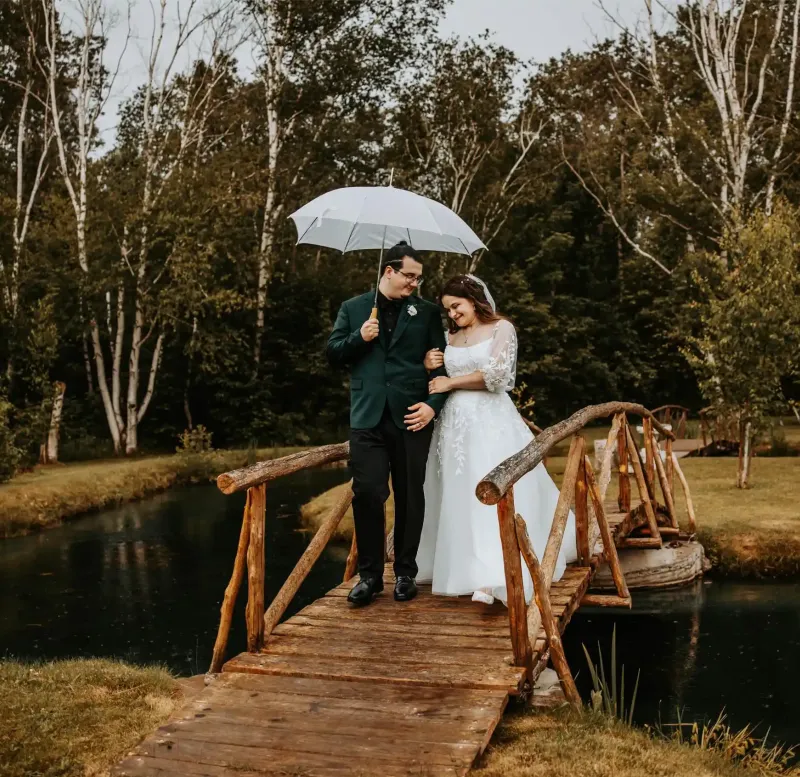 A bride and groom on a wooden bridge with an umbrella on a cloudy day at Castle Farms.