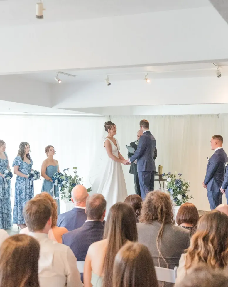 A bride and groom with a wedding party and guests at a wedding in a white-walled gallery at Castle Farms. 