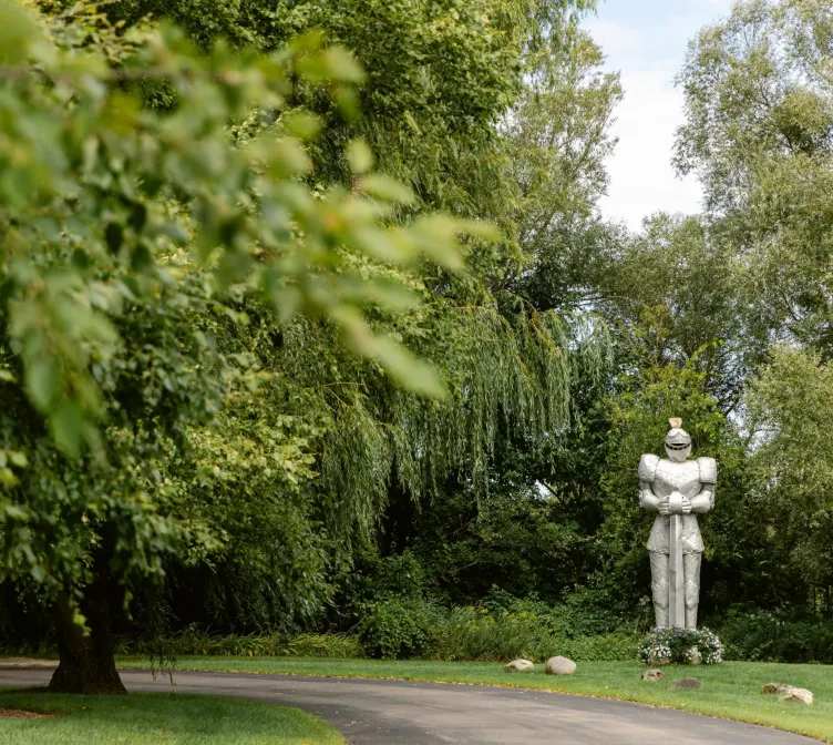 A knight in shining armor standing in a grove of green trees beside a road at Castle Farms.