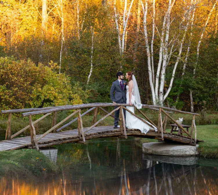 A bride and groom on a wooden bridge over water with trees at sunset in autumn at Castle Farms.