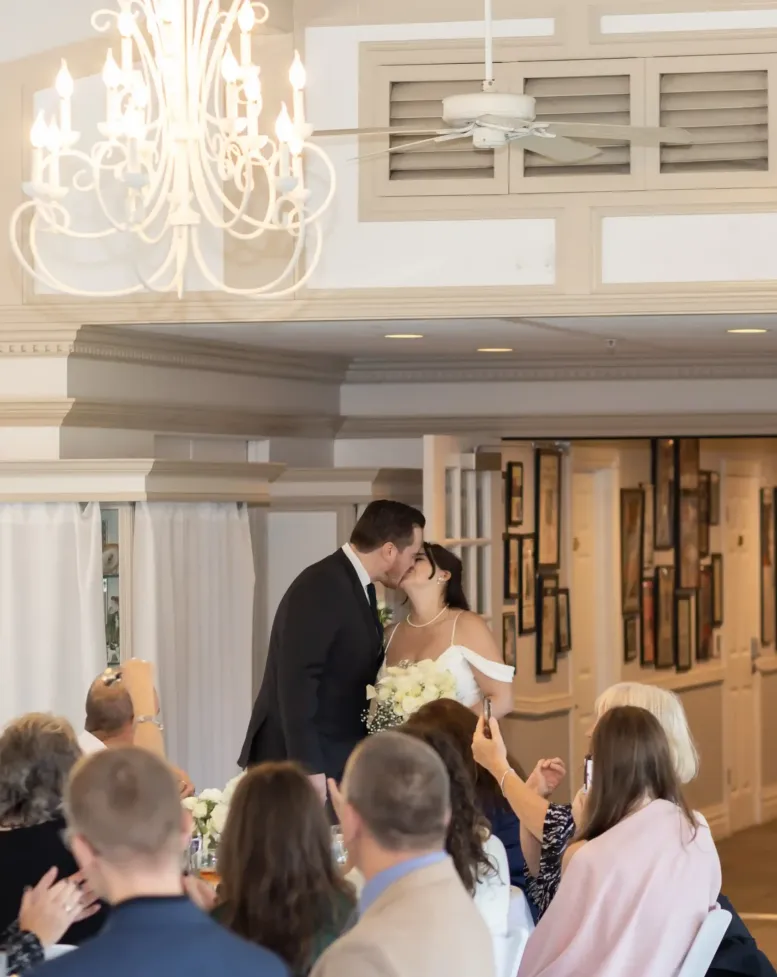 A bride and groom kissing in front of guests in a reception space with ornate chandeliers at Castle Farms.