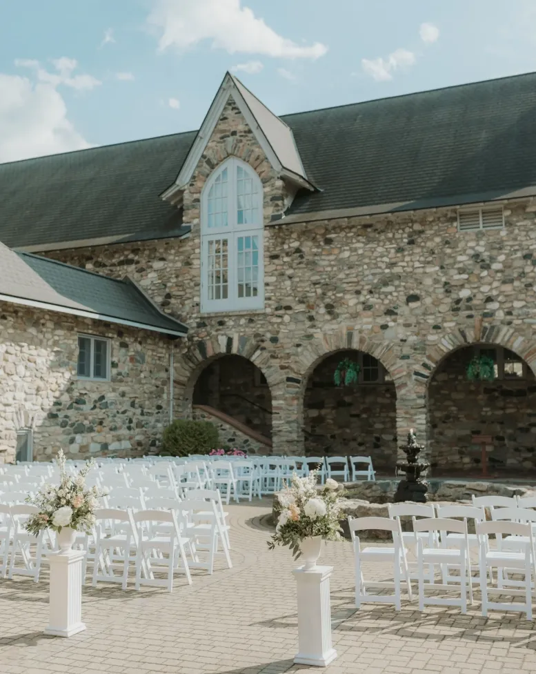 Rows of white chairs and white flower stands in a stone courtyard next to a building with a window at Castle Farms.